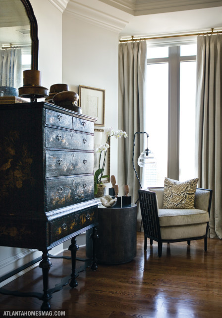Living room corner with dark lacquered chest, armchair with patterned pillow, and tall windows with beige curtains.