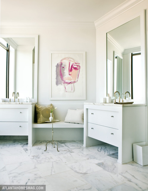 White bathroom with marble tile floors, two white vanities with brass faucets, and a built-in bench with abstract art above