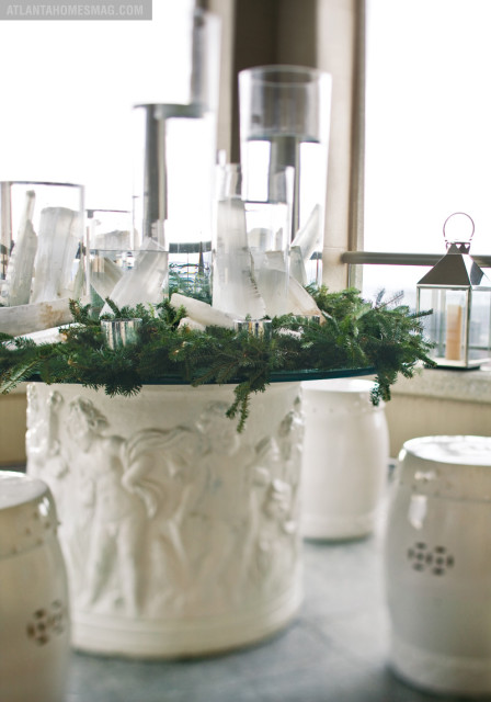 White pedestal with bas-relief figures, topped with a glass surface displaying a festive arrangement of evergreen branches, clear glass cylinders, and white crystal shards.