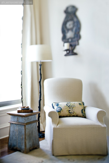 Cream upholstered armchair with a floral lumbar pillow sits next to a distressed blue side table and a floor lamp with a branch-like stem.