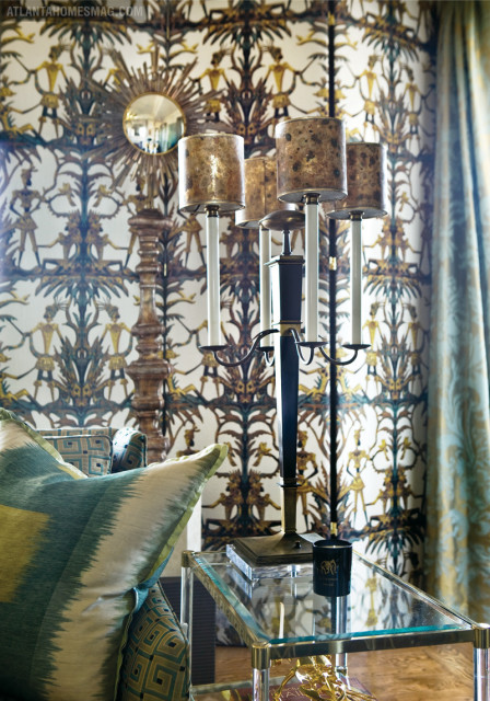 Living room with bold patterned wallpaper, a gold sunburst mirror, a tall candelabra, and a sofa with teal and green ikat pillows.