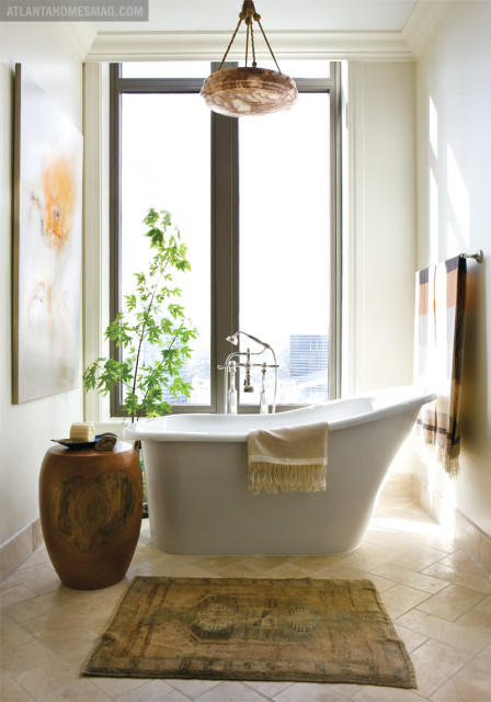 Freestanding white bathtub with chrome fixtures, a beige towel draped over the side, and a plant in a wooden stool beside it. A patterned rug lies on the tiled floor in front of the tub.