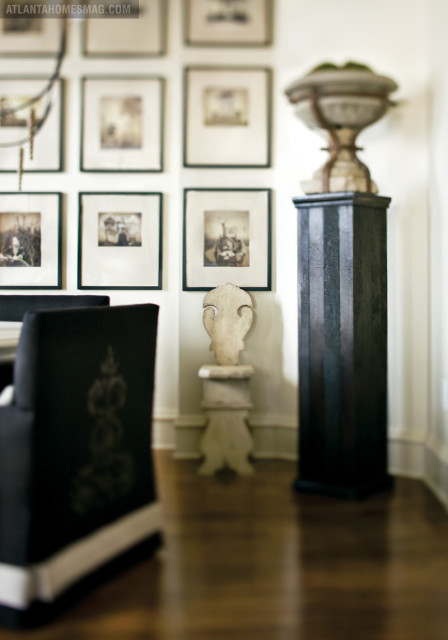 Dining room with dark upholstered chairs, a gallery wall of framed vintage photographs, and a tall black pedestal topped with a stone urn.