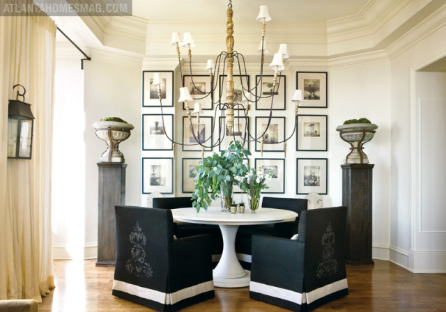 Dining room with a round white pedestal table surrounded by four black upholstered chairs with white trim and ornate detailing. A large, ornate chandelier hangs above. The wall is adorned with a grid of framed black and white prints.