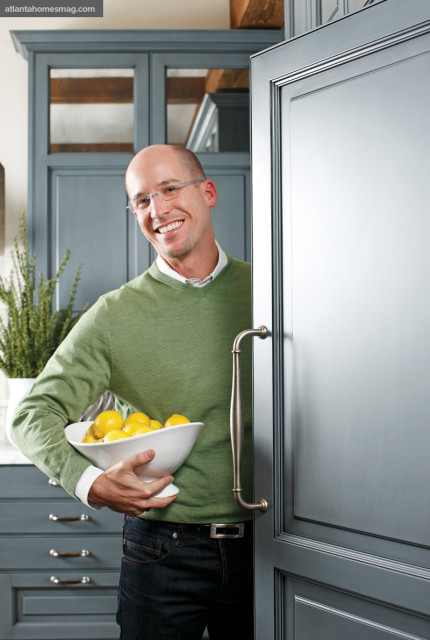 Man in a green sweater holding a bowl of lemons in a gray kitchen with custom cabinetry and wood beams.