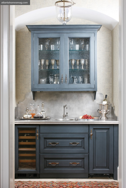 Blue kitchen cabinetry with glass-fronted upper cabinets displaying glassware and silver, a marble countertop with a sink, and a wine fridge.