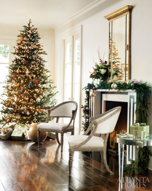 Living room decorated for Christmas with a lit tree, fireplace garland, two antique chairs, and gifts on a dark wood floor.