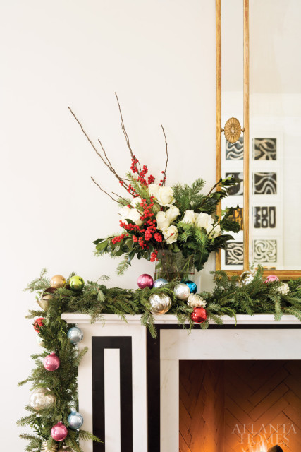Marble fireplace decorated for Christmas with a garland of fir branches, ornaments, and a floral arrangement of white roses and red berries.