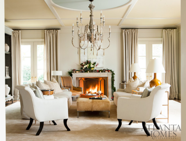Living room with a fireplace decorated for Christmas, two white armchairs, a sofa, and a cowhide ottoman. Neutral tones and a large chandelier dominate the space.