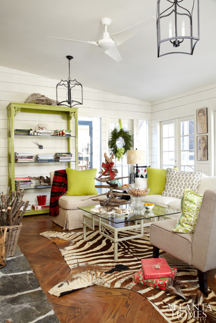 Living room with white shiplap walls, a white ceiling fan, and a black lantern chandelier. Features a lime green bookshelf, a tan armchair, a sofa with lime green pillows, a glass coffee table with a zebra rug, and a fireplace with stacked logs.