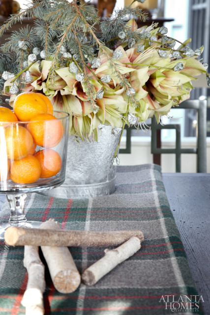 Close-up of a floral arrangement with light green and pink amaryllis flowers, pine branches, and small white berries in a galvanized bucket, next to a glass bowl of tangerines on a plaid runner.