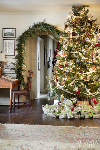 Festive living room with a decorated Christmas tree, gifts wrapped in silver and green, and a garland draped over an arched doorway. A wooden desk and chair are visible to the left.