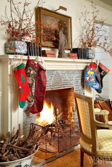 Stone fireplace with a roaring fire, decorated for Christmas with stockings hanging from the mantel. A painting hangs above the mantel, flanked by vases of red berries and branches.