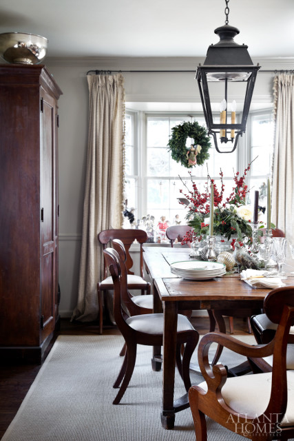 Dining room with wooden table set for a holiday meal, featuring a centerpiece of red berries and greenery, Christmas wreaths on the window, and a large dark wood cabinet.