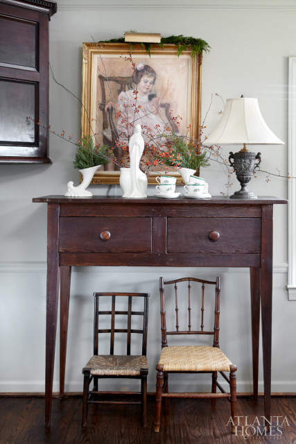 Dark wood console table with two drawers topped with a painting of a young girl, decorative vases, teacups, and a lamp. Two antique chairs sit below.