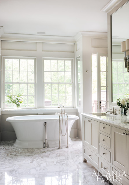 White bathroom with freestanding tub, marble floors, and a vanity with white cabinetry and marble countertop. Large windows overlook greenery.