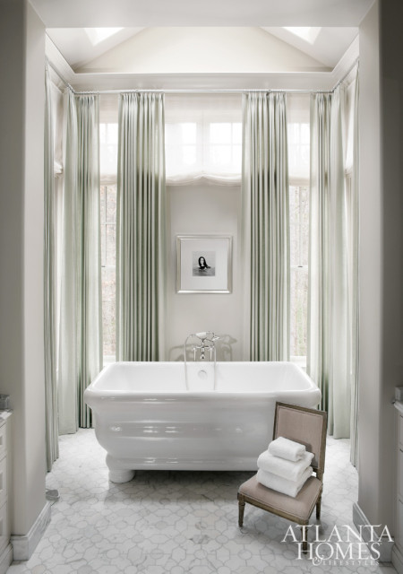 White freestanding bathtub in a bathroom with pale green floor-to-ceiling curtains, marble tile floor, and a framed black and white photograph above the tub.