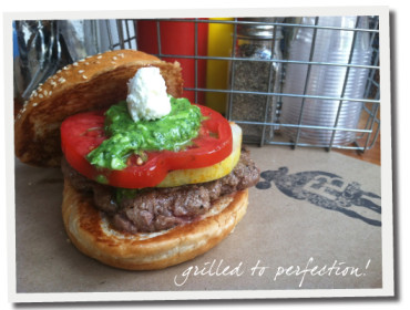 Burger with sesame seed bun, beef patty, tomato, green sauce, and white cheese, with condiments in the background.