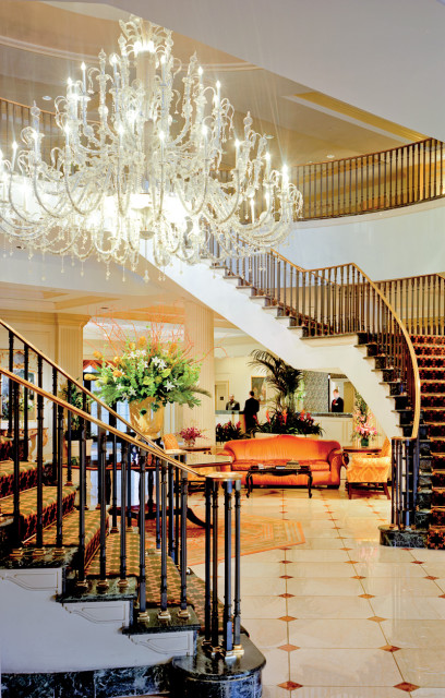 Grand foyer with a large crystal chandelier, a curved staircase with patterned carpet, marble floors, and an orange sofa