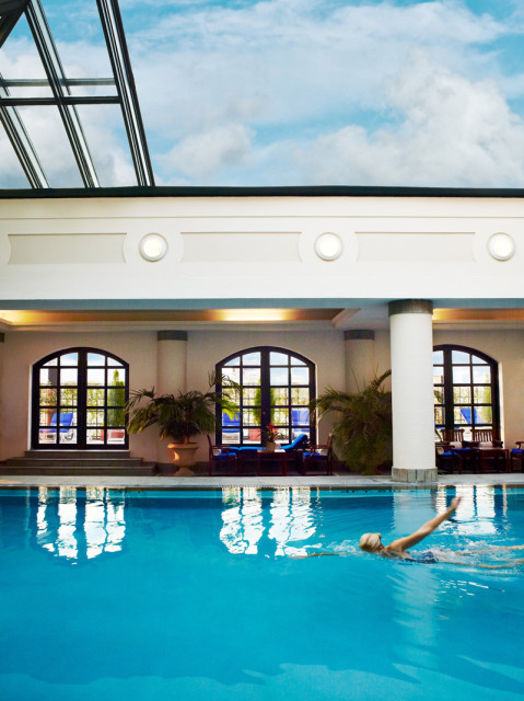 Indoor swimming pool with bright blue water, a person swimming, arched windows with dark frames, and a glass ceiling