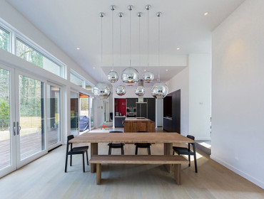 Modern dining room with light wood floors, a long wooden dining table with black chairs and a bench, and a cluster of large, reflective spherical pendant lights overhead. Large windows and sliding glass doors overlook a deck and wooded area.