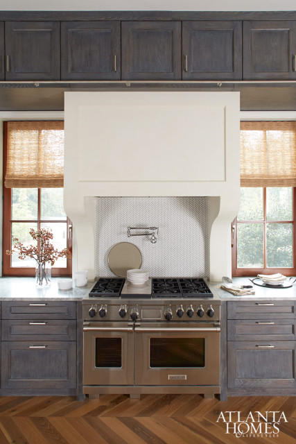 Dark wood kitchen cabinets flank a stainless steel range with a white hood and herringbone tile backsplash. Woven blinds cover windows on either side.