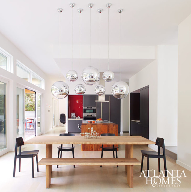 Modern dining room and kitchen with a light wood table, black chairs, and a bench. Above the table hang multiple large, spherical chrome pendant lights. The kitchen features dark cabinetry, a red backsplash, and a wood island.