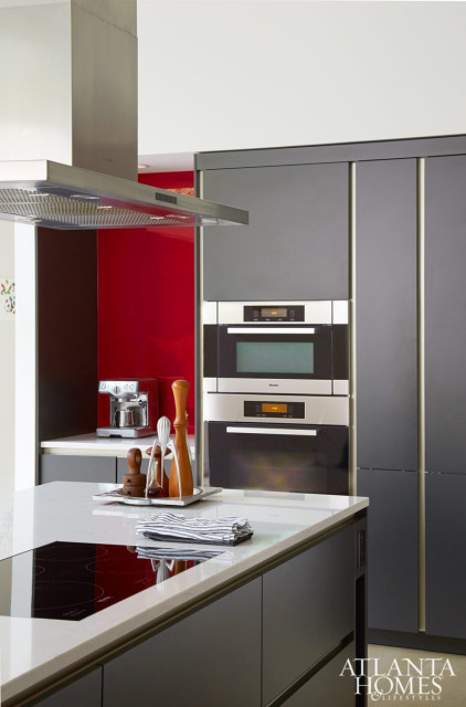 Modern gray kitchen with a red backsplash, stainless steel appliances including a coffee maker and stacked ovens, and a white island with an induction cooktop.