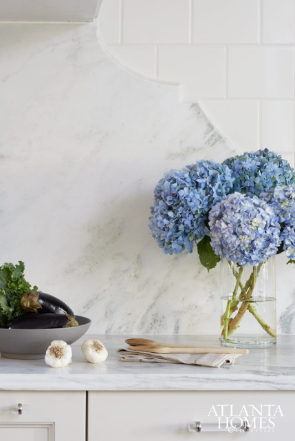 White marble backsplash with a decorative curve above a white subway tile backsplash in a kitchen. A vase of blue hydrangeas sits on a marble countertop next to a bowl of eggplants and garlic.