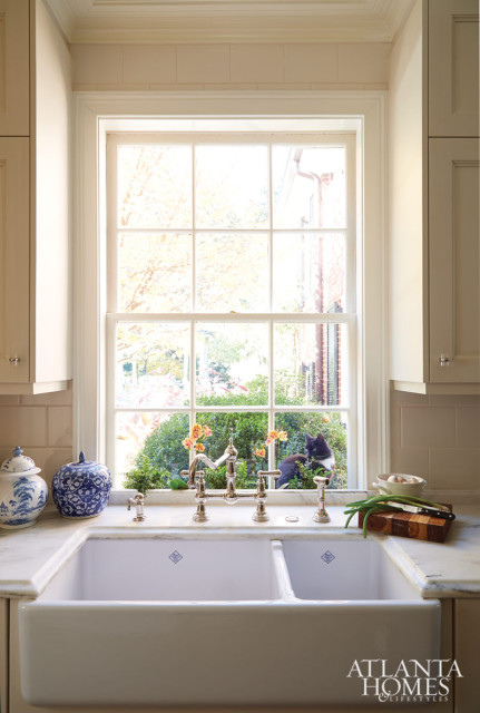 White farmhouse sink with marble countertops and a bridge faucet, set against a window overlooking a garden with a black cat.