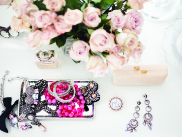 Silver tray with assorted jewelry including pink beaded necklaces, diamond bracelets, and a black ribbon bow. A perfume bottle, clutch, and earrings are nearby.
