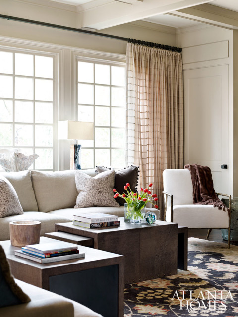 Living room with a sectional sofa, white armchair, dark patterned rug, and a large window with sheer curtains. A dark wood coffee table holds books and a vase of red tulips.