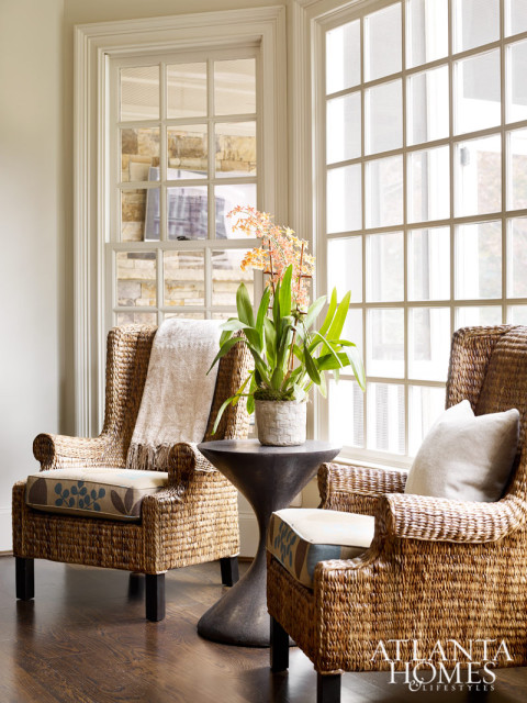 Two wicker armchairs with patterned cushions flank a pedestal side table holding an orchid plant, with large grid-pane windows in the background.