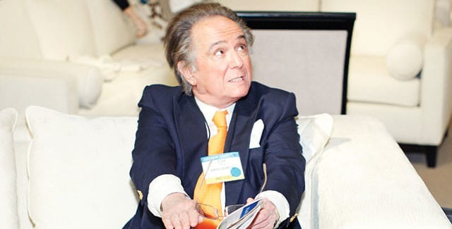Man in a navy suit and orange tie wearing a name tag, holding glasses and papers.