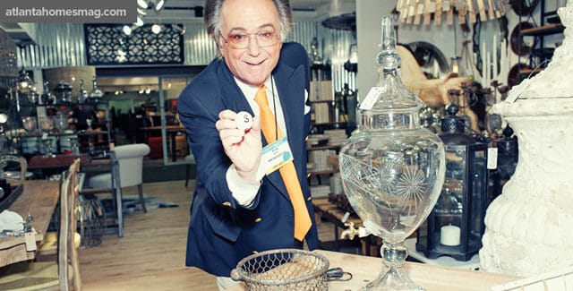 Man in a navy blazer and orange tie holds up a lottery ball with the letter S, standing in a store filled with decorative items.