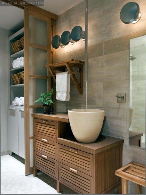 Bathroom vanity with slatted wood drawers and a cream vessel sink, next to a closet with shelves of towels and baskets.