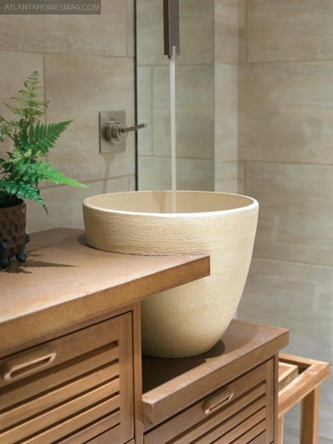Beige vessel sink with textured finish on a wood vanity with slatted drawers. Water flows from a brushed nickel faucet against a tiled wall.