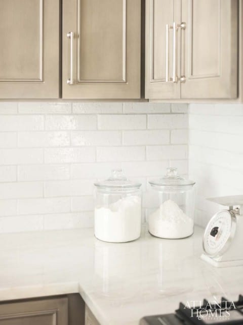 Kitchen with taupe shaker cabinets, white subway tile backsplash, marble countertops, glass jars of flour, and a vintage scale.