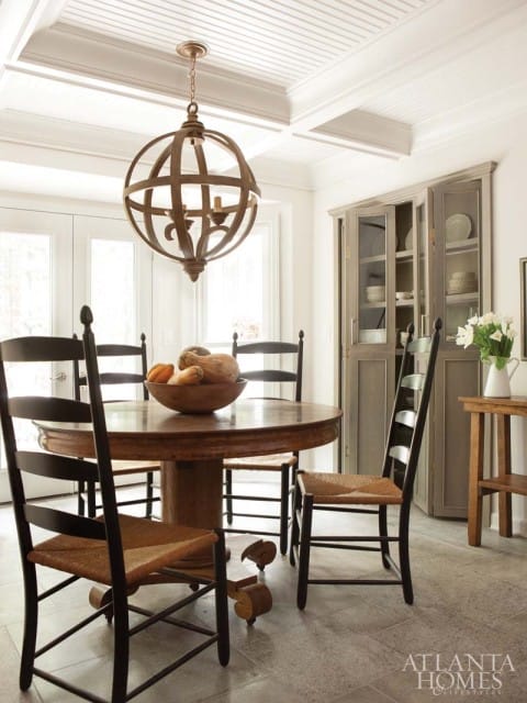 Round wooden dining table with woven chairs and a large globe chandelier in a room with a gray hutch and tiled floor.