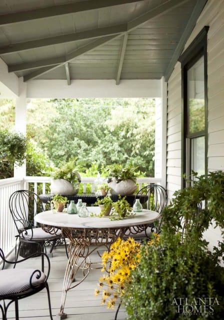 Covered porch with a marble-top table and chairs, surrounded by potted plants and yellow flowers.