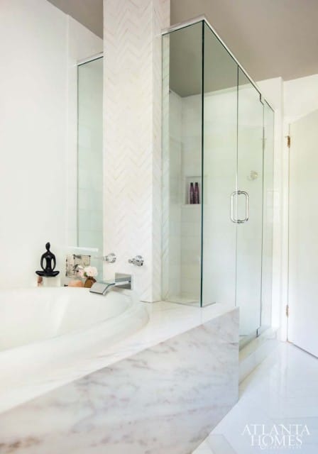 White bathroom with a soaking tub and a glass-enclosed shower, featuring marble countertops and herringbone tile accents.