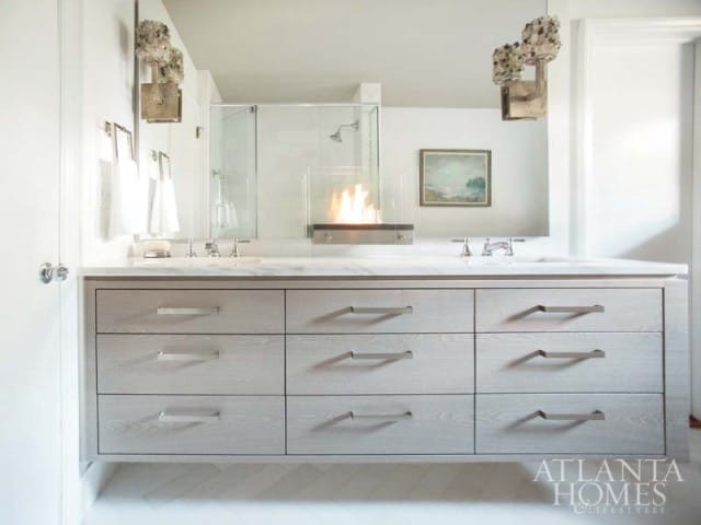 Modern bathroom with a double vanity featuring light wood drawers, marble countertop, and chrome fixtures. A glass-enclosed fireplace is visible behind the sinks.