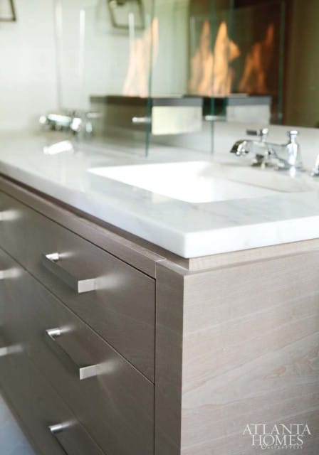Modern bathroom vanity with light wood drawers, marble countertop, undermount sink, chrome faucet, and a glass-enclosed fireplace with flames.
