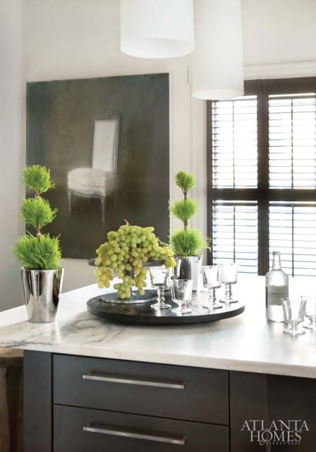 Kitchen island with marble countertop, dark gray cabinetry, and a tray with grapes, topiary plants, and glassware. A painting of a chair hangs on the wall.