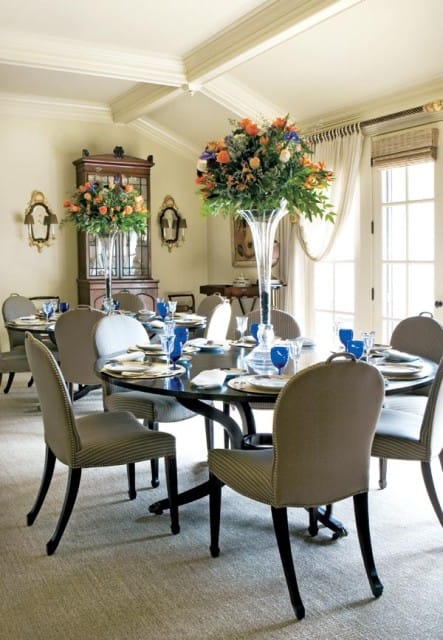 Formal dining room with a round dark wood table set for eight with blue glassware, neutral striped chairs, and large floral arrangements in glass vases.