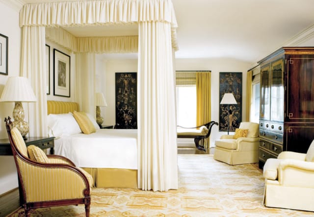 Bedroom with a four-poster bed draped in cream fabric, yellow striped armchair, cream upholstered chairs, and a dark wood armoire with brass accents.