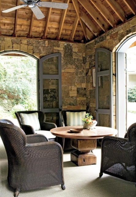 Covered patio with stone walls, wood-beamed ceiling, and ceiling fan. Wicker chairs surround a round wooden table with a floral centerpiece.