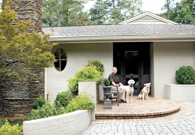 Man sitting on a bench on a brick patio with two dogs in front of a light-colored brick house with a stone chimney and an oval window.