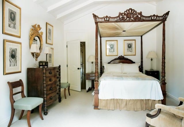 Bedroom with white walls, vaulted ceiling, and a dark wood four-poster bed. Antique dresser with gold mirror and framed portraits on the wall.