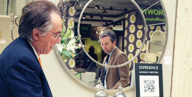 Man in navy suit and orange tie looks into a round mirror reflecting another man in a brown suit. A sign reads "EXPERIENCE MODERN HISTORY" with a QR code.